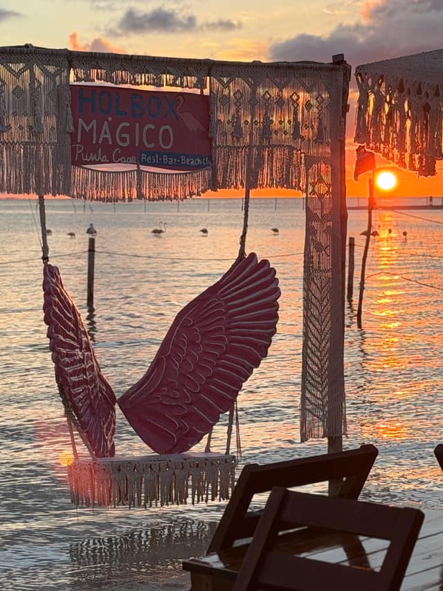 El sol toca el horizonte a través del marco de macramé con alas y el letrero de Holbox Mágico